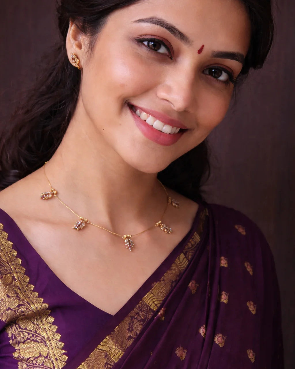 Woman wearing a purple saree with gold patterns and jewelry against a brown background