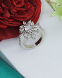 Silver ring with clear gemstones on a white surface with a red flower and green leaves in the background