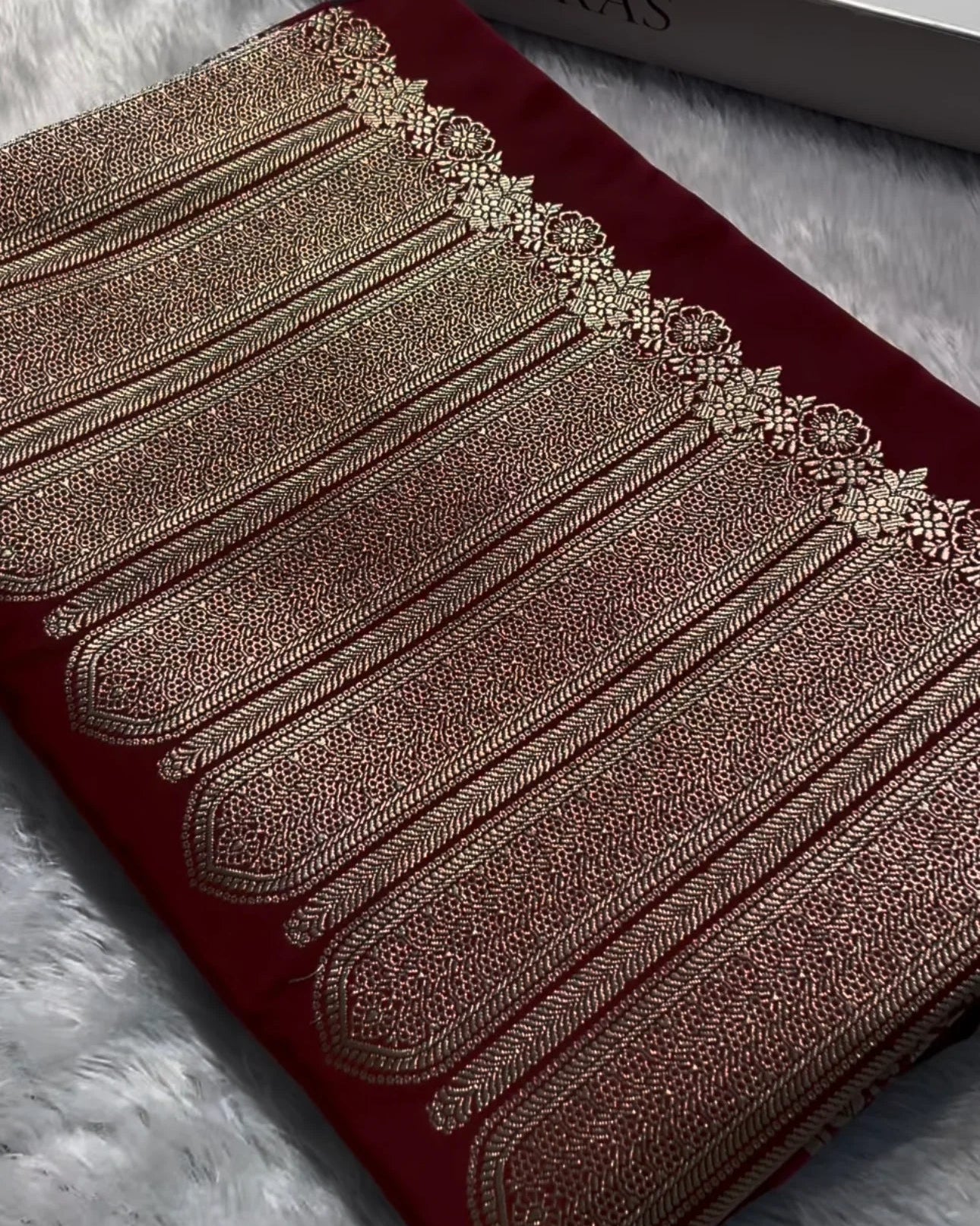 a folded deep red fabric garment with ornate metallic gold embroidery and a decorative border, placed on a white faux-fur surface next to a white banaras box.