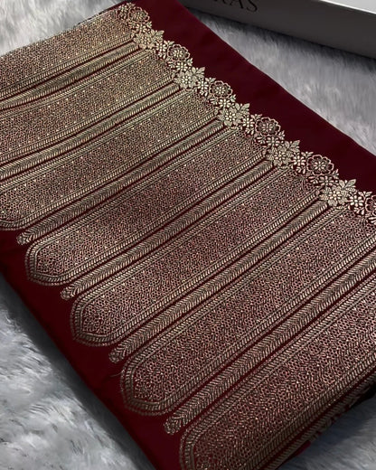 A folded deep red fabric garment with ornate metallic gold embroidery and a decorative border, placed on a white faux-fur surface next to a white Banaras box.