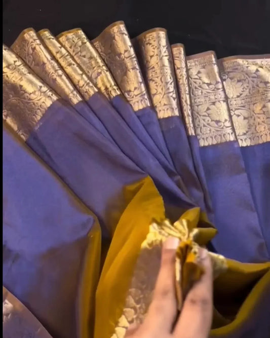 A close-up of a purple and gold saree being held open by a hand, showing its metallic zari border and yellow-gold inner layer against a dark background.