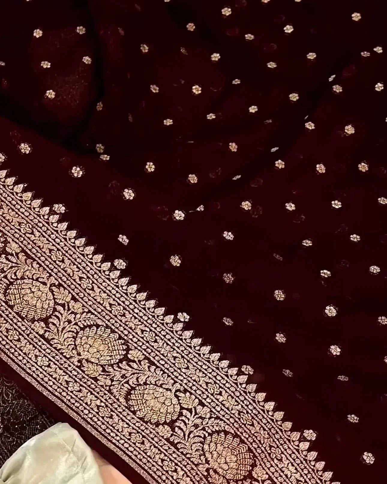 a close-up of a deep maroon velvet fabric with intricate gold zari embroidery, featuring small scattered motifs and a wide ornate border. a hand wearing a white sleeve is holding the fabric to show its drape and texture.