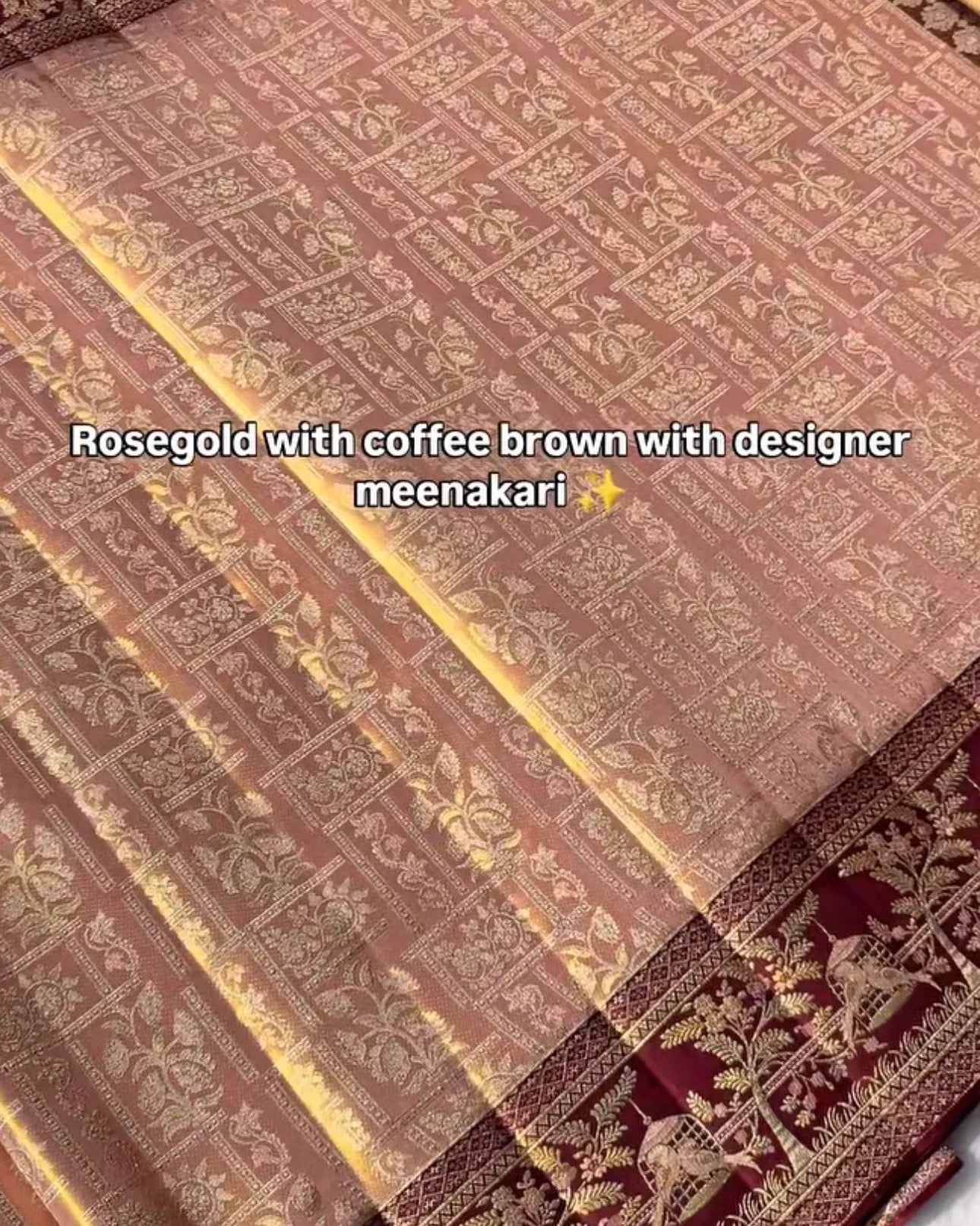 A close-up of a folded and draped fabric, likely a saree or scarf, featuring a rosegold and coffee brown base with intricate gold floral patterns and a deep red border with gold motifs. White text overlay reads 'Rosegold with coffee brown with designer meenakari'.