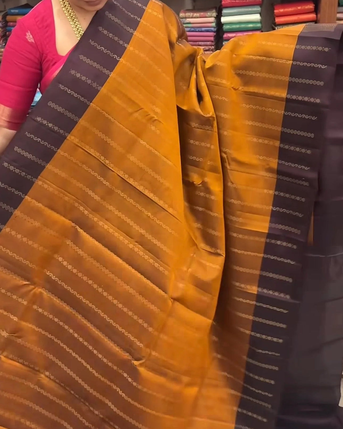 a woman in a pink top holds up a large mustard yellow and black saree with metallic zari stripes, with shelves of colorful folded fabrics in the background.