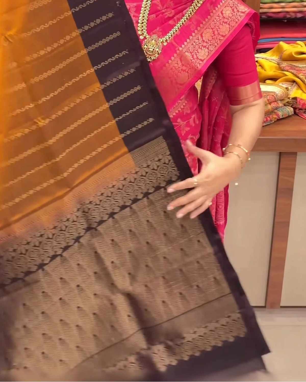 A woman in a pink saree holds up a black and gold Banarasi silk saree to display its intricate zari work and border pattern in a fabric store.