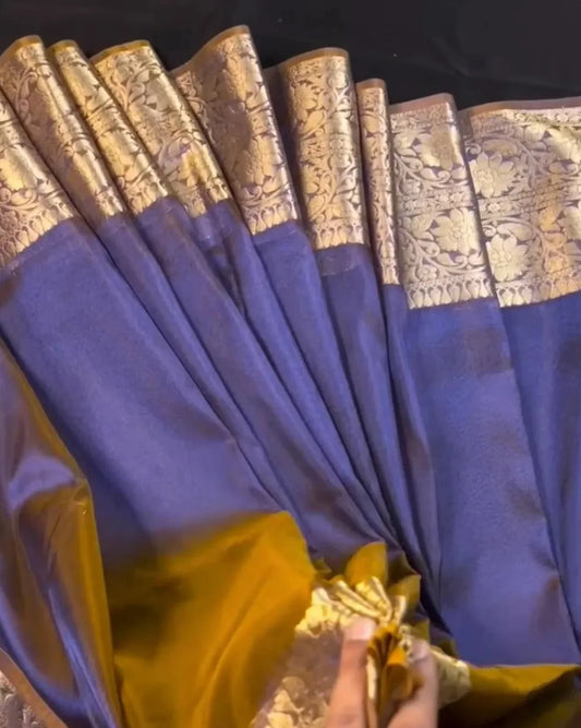 A close-up of a blue and gold silk saree being held by a hand, showing the rich texture and metallic zari embroidery.