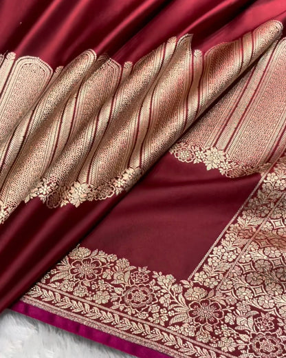 A close-up of a richly colored silk saree, showing a deep maroon body with intricate gold zari Banarasi borders and a contrasting metallic gold body section, laid over a white faux fur surface.