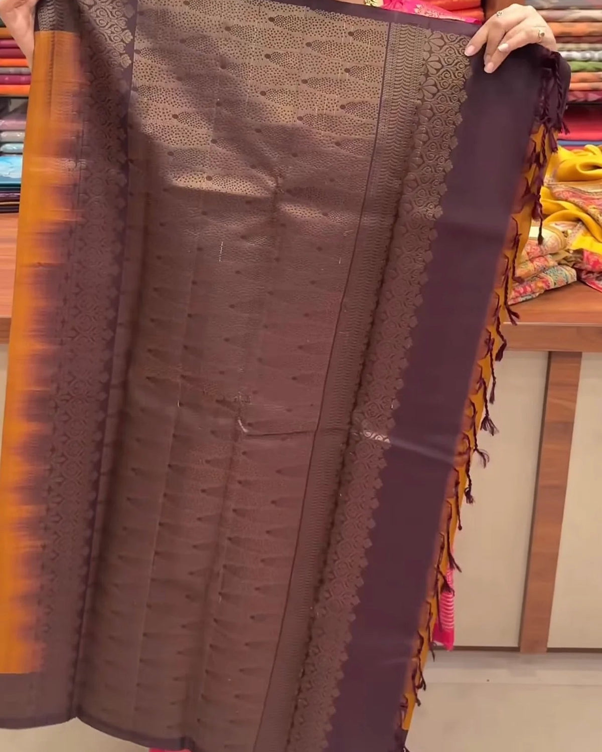 A person holds up a dark brown and purple silk saree with gold zari embroidery and orange border accents in a fabric store.