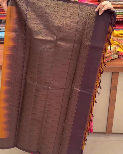 A person holds up a dark brown and purple silk saree with gold zari embroidery and orange border accents in a fabric store.