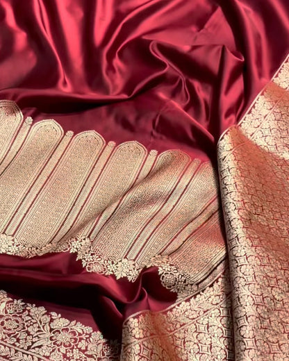 A close-up of a richly textured red and gold fabric, likely a silk saree or scarf, featuring a deep red body with intricate gold zari brocade patterns and a scalloped border.
