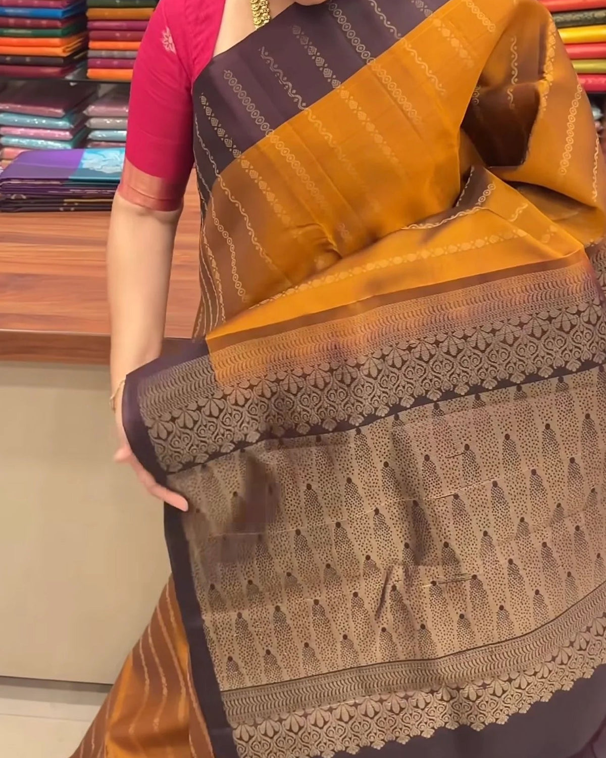 A woman wearing a traditional Indian saree in a fabric store. The saree features a mustard yellow and dark brown body with intricate woven patterns, paired with a bright pink blouse.