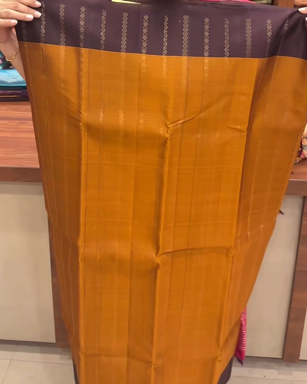 a woman in a pink top holds up a wide orange-gold fabric saree or sari with a dark purple border and metallic vertical motifs, photographed inside what appears to be a fabric or apparel store.