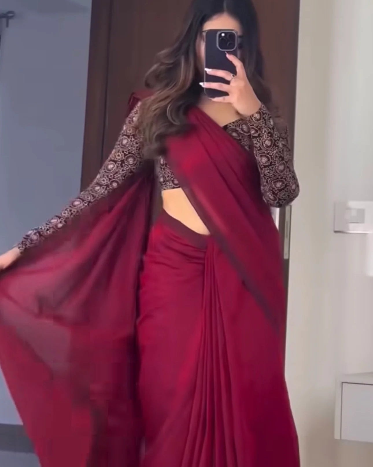 a woman takes a mirror selfie wearing a deep red sari with sheer black patterned sleeves and a side cutout, holding out one side of the drape.