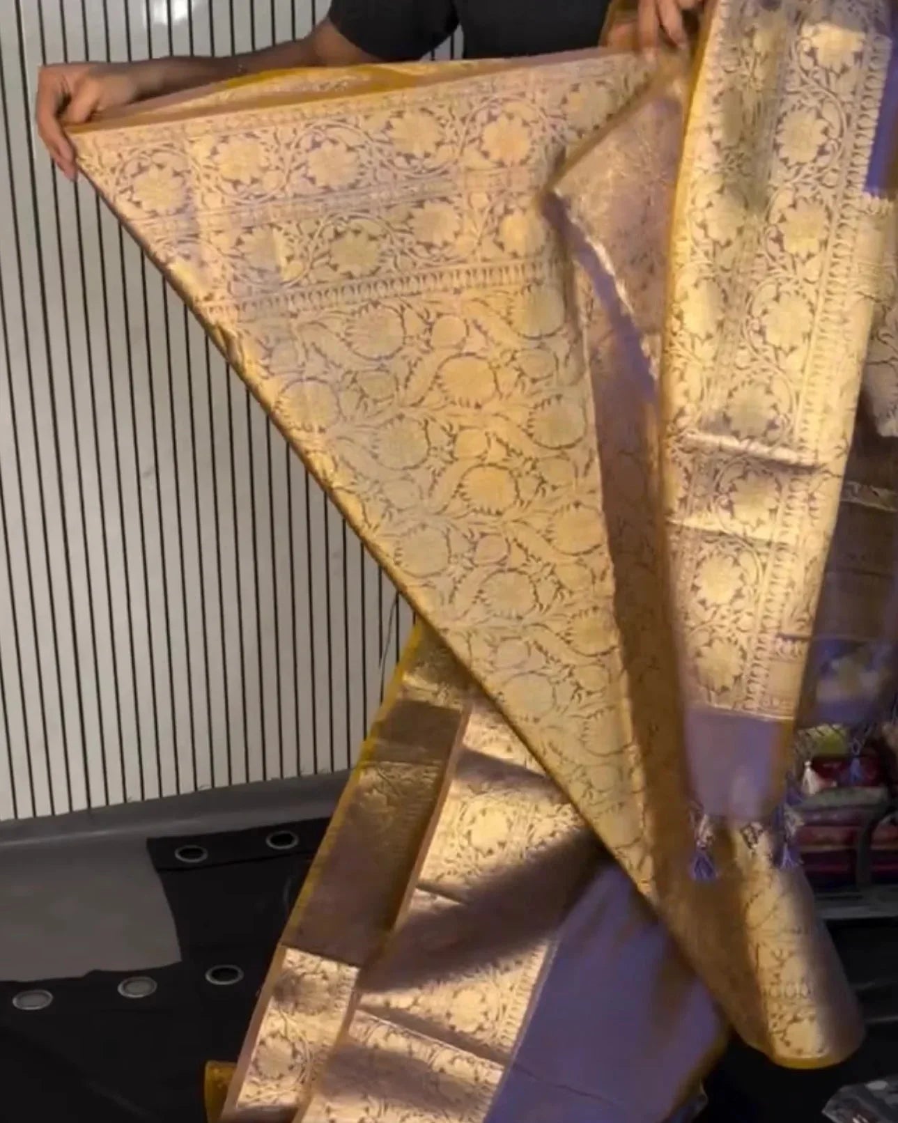 a person holds up a folded and draped silk saree with intricate gold zari floral patterns on a lavender-purple base, set against a striped wall background.