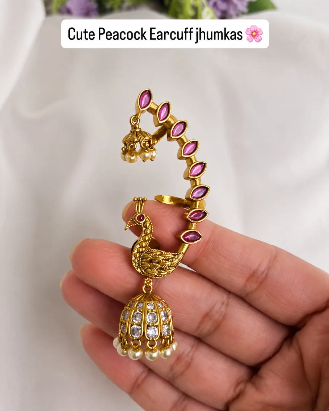 Gold peacock earrings with pink stones held by a hand against a white background
