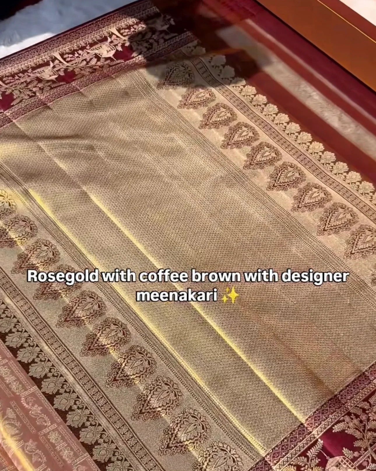 A close-up of a traditional Indian saree with a beige body, rose gold and gold zari borders, and a deep red pallu. Text overlay reads 'Rosegold with coffee brown with designer meenakari'.